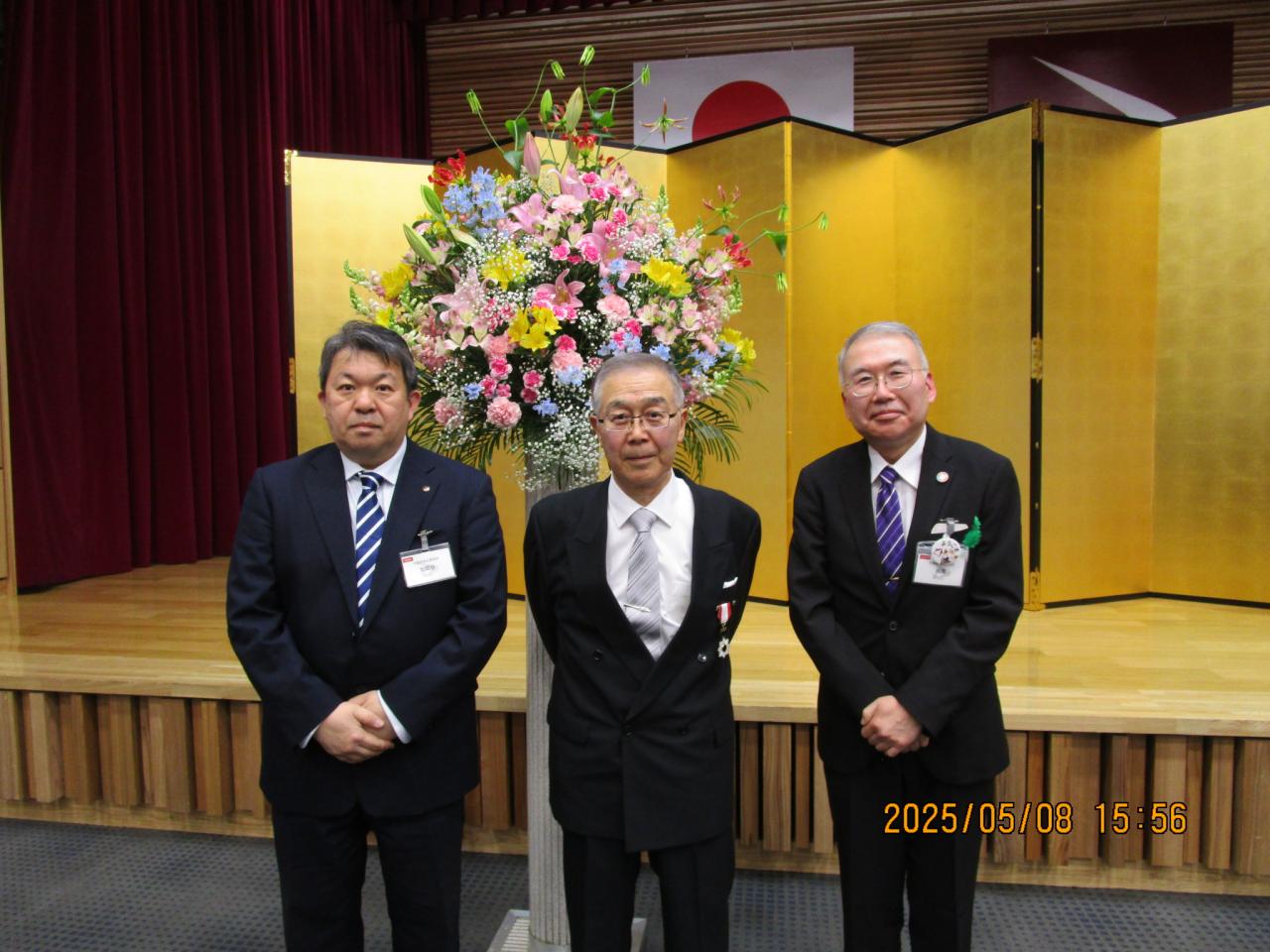 秋田県正庁において、金屏風と花の前で、叙勲受章者を真ん中に、両脇を秋田県労働員会事務局長と審査調整課長が並んでいる写真